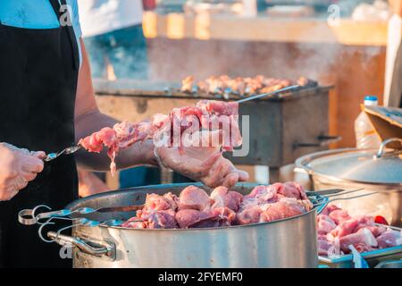 Die Hände eines Mannes, der Fleisch auf einem Grillspieß spieß. Nahaufnahme von rohem Fleisch auf einem Spieß. Street Food Festival. Stockfoto