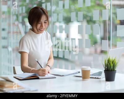 Eine kurze Aufnahme einer Studentin, die Aufgaben macht und auf einem Notizbuch in einer modernen Bibliothek schreibt Stockfoto