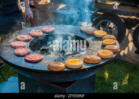 Auf einem großen, runden Holzgrill draußen werden Rindersteaks und Burger-Brötchen gegrillt. Grillfest im Stadtpark. Street Fast Food. Stockfoto