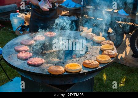 Auf einem großen, runden Holzgrill draußen werden Rindersteaks und Burger-Brötchen gegrillt. Grillfest im Stadtpark. Street Fast Food. Stockfoto