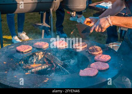 Auf dem Rasen röstet ein großer runder Holzgrill Rindersteaks für Burger. Grillfest im Stadtpark. Street Fast Food. Stockfoto