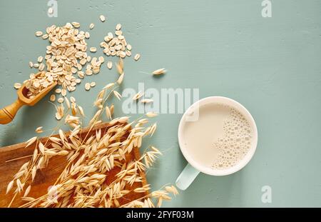 Hafermilch im Glas und Becher auf blauem Hintergrund. Flocken und Ohren für Haferflocken und Müsli auf einem Holzteller. Stockfoto