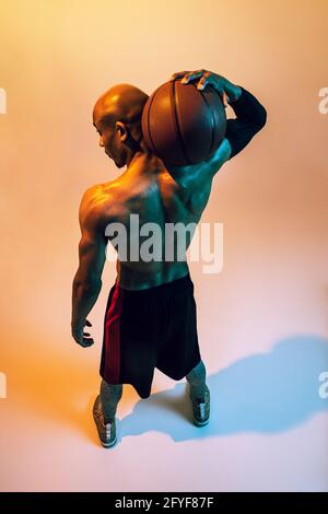 Sportlicher, muskulöser afroamerikanischer Basketballspieler, der in Neonlicht auf orangefarbenem Hintergrund posiert. Rückansicht Stockfoto