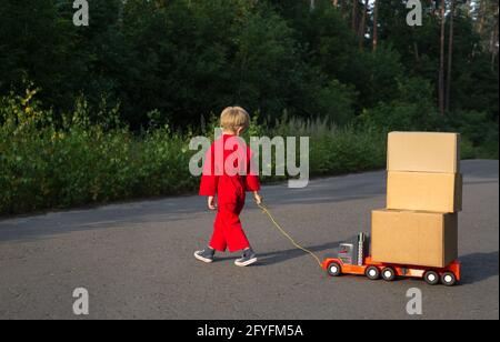 Kind in einem roten Overall geht die Straße entlang und trägt ein großes Spielzeugauto - einen Lastwagen mit Kartons darauf. Kleiner Kurier bei der Arbeit. Paketzustellung, Stockfoto