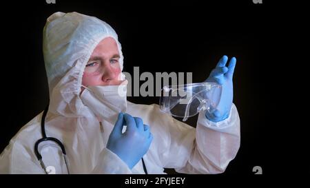 Ein Sanitäter in einem weißen Schutzanzug mit Schnitten aus einer Maske und Brille auf seinem Gesicht, schwarzer Hintergrund Stockfoto