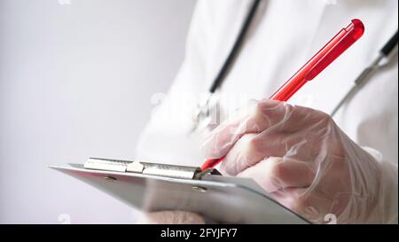 Nahaufnahme eines Arztes, der ein Rezept in einer Krankenakte auf eine Tablette schreibt. Ein Arzt in einem weißen Mantel und Handschuhen schreibt ein Rezept mit einem roten Stockfoto