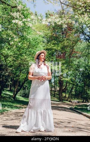 Ganzkörperportrait einer Frau mittleren Alters, die im blühenden Frühlingspark spazieren geht. Stilvolle Dame trägt Hut und weißes Kleid im Freien unter Magnolienbaum Stockfoto