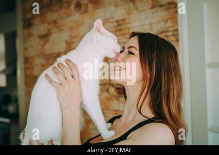 Lächelnde Frau mit einer weißen Katze, die ihr Gesicht leckt Stockfoto