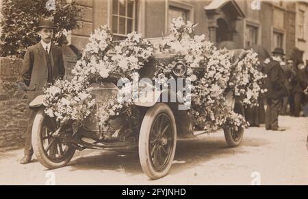 Eine alte Fotopostkarte aus dem frühen 20. Jahrhundert, die einen Mann zeigt, der neben einem mit Blumen bedeckten Auto steht. Verkleidet für eine Hochzeit oder Beerdigung. Stockfoto