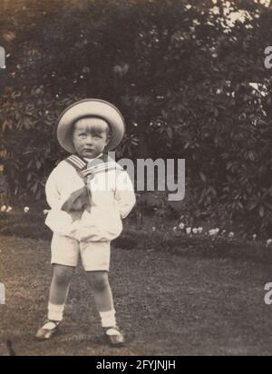 Eine alte Fotopostkarte aus dem frühen 20. Jahrhundert, auf der ein kleines Kind in einem Garten mit Matrosenoutfit stand. Stockfoto