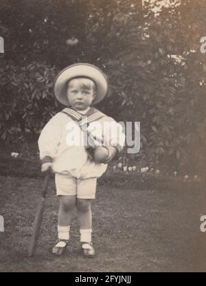 Eine alte Fotopostkarte aus dem frühen 20. Jahrhundert, auf der ein kleines Kind in einem Garten mit Matrosenoutfit stand. Halten Sie einen Cricket-Schläger und Ball. Stockfoto