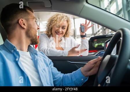 Junges Paar, das ein neues Auto kauft. Frau zeigt ihrem Mann, der im Autohaus sitzt, den Schlüssel zum neuen Auto Stockfoto