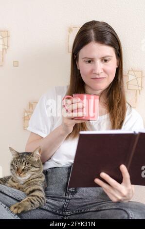 Eine junge Frau im Alter von 30 Jahren in einem weißen T-Shirt liest ein Buch und hält eine Tasse Kaffee in der Hand. Die gestromte Katze liegt auf dem Bein des Besitzers. Heimkomfo Stockfoto
