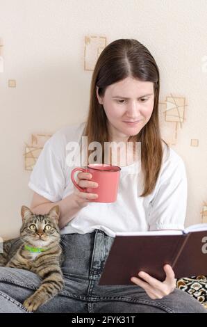 Eine junge Frau im Alter von 30 Jahren in einem weißen T-Shirt liest ein Buch und hält eine Tasse Kaffee in der Hand. Die gestromte Katze liegt auf dem Bein des Besitzers. Heimkomfo Stockfoto