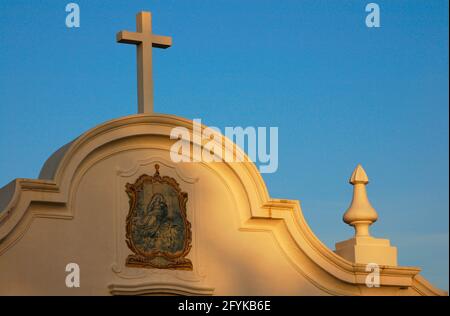 Portugal, Sines. Kirche Nossa Senhora das Salas. Die Kapelle wurde auf Initiative von Vasco da Gama erbaut, neben dem Ort, an dem sich eine Einsiedelei befand, die an die Rettung der Prinzessin Fatassa erinnert. Der ursprüngliche Tempel stammt aus dem 15. Jahrhundert, obwohl er im 16. Jahrhundert modifiziert wurde. Architektonisches Detail des oberen Teils der Hauptfassade, mit einem Kreuz und einer kegelförmigen Spitze, die die Strebepfeiler der Kirche krönt. Bezirk Setúbal. Stockfoto