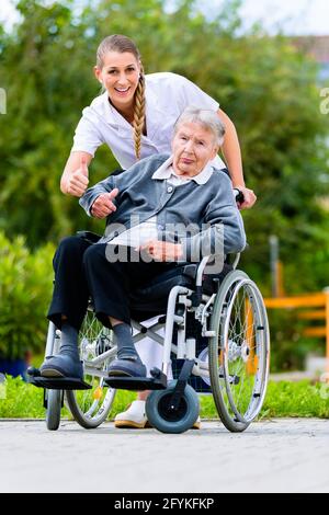 Ältere Frau im Pflegeheim mit Krankenschwester im Garten sitzen im Rollstuhl, den Daumen nach oben anmelden Stockfoto