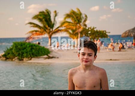 Süßer kaukasischer Teenager-Junge mit nassen Haaren, die abends vor einer verschwommenen kleinen sandigen Insel mit Palmen, Touristen und Strandplankenbetten posieren. Alles Stockfoto