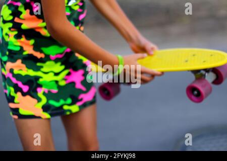 Defokussieren Mädchen mit gelben Penny Board. Vorderansicht. Jugend-Hipster-Kultur. Kinderhände halten tagsüber ein kurzes Cruiser-Pennyboard im Freien. Aktive Kinder Stockfoto