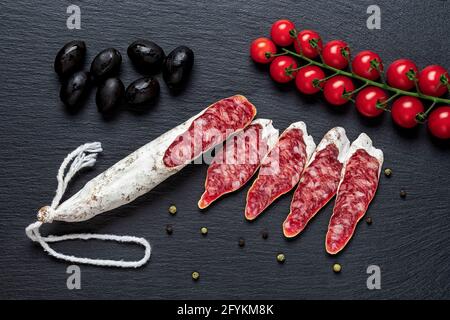 Scheiben von fetten katalanischen dünnen Würstchen, schwarzen Oliven und Kirschtomaten auf der Weinrebe über schwarzer Schieferfläche. Traditionelle spanische, trocken gehäutete Schweinefleischsalami. Stockfoto