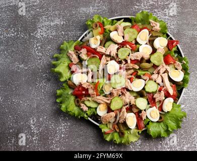Nicoise Salat mit Thunfisch, Gurken, Paprika, Zwiebeln, Wachteleiern, Bohnen und Kartoffeln auf einem runden Teller auf dunklem Grund. Draufsicht, flach liegend Stockfoto
