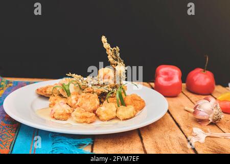 Peruanische Küche: Sautierte Garnelen mit Kräutern und Knoblauch, Holztisch, serviert auf einem weißen Teller, begleitet von purpurem Maissaft Stockfoto