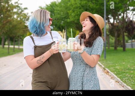 Zwei glückliche Freundinnen trinken erfrischende Getränke, lachen und schauen sich an einem sonnigen Sommertag in einem Park an. Stockfoto