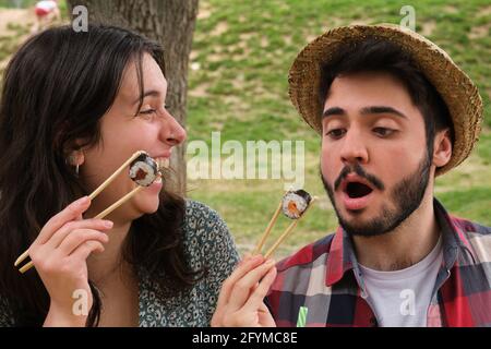Ein junges, glückliches Paar lacht und isst Sushi in einem Park, wo es an einem sonnigen Sommertag ein Picknick macht. Stockfoto