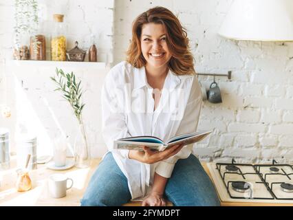Wunderschöne glückliche junge Frau plus Größe Körper positiv in blau Jeans und weißes Hemd lesen Kochbuch in der heimischen Küche Stockfoto