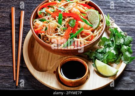 Nahaufnahme von Pad thai Nudeln mit Garnelen, Karotten, rotem Pfeffer, Koriander und Limette in einer Schüssel auf einem Bambusblech mit Sojasauce und Essstäbchen Stockfoto