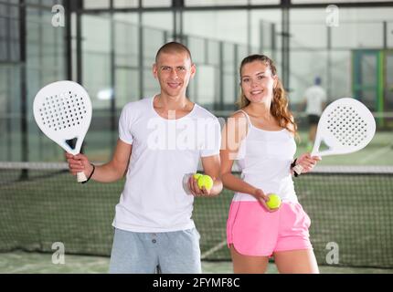 Porträt einer lächelnden jungen Frau und eines sportlichen Mannes, der in der Halle auf dem Padel-Platz mit Schlägern und Tennisbällen posiert Stockfoto