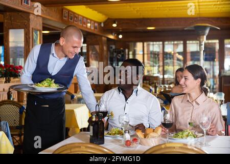 Der Kellner servierte Mahlzeiten für ein positives gemischtradetes Paar im Restaurant und stellte Teller mit Essen auf den Tisch Stockfoto