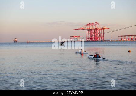 Zwei Ruderer in einem Kajak im ruhigen Meerwasser und die im Hintergrund aufstellenden Frachtschräne im Hafen. Stockfoto