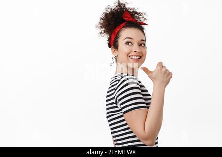 Porträt einer jungen Frau, einer Frauenrechtlerin, die mit dem Finger nach links auf ihre Schulter zeigt, lächelnd und hoffnungsvoll aussagt und gegen Weiß steht Stockfoto