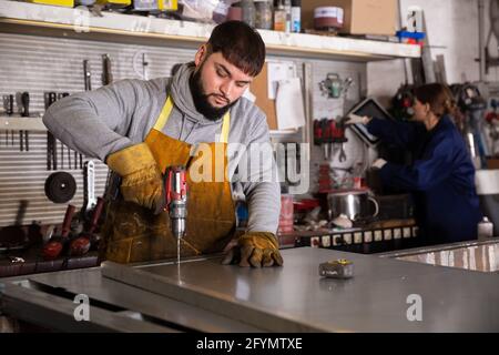 Seriöse junge Handwerker arbeiten mit manuellen elektrischen Bohrer, Bohren von Metallstrukturen in der industriellen Werkstatt Stockfoto