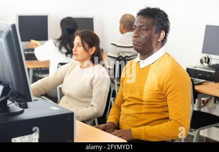 Fokussierter junger erwachsener Mann, der mit Computer und Büchern am Tisch sitzt und in der Bibliothek Informationen findet Stockfoto