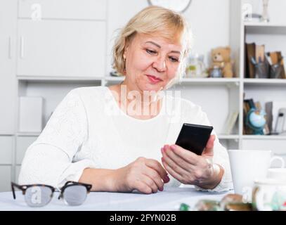 Reife Frau am Tisch zu Hause sitzen und auf Smartphone in den Händen Stockfoto