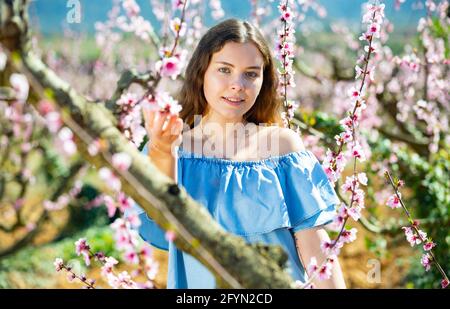 Positive europäische Mädchen in blauem Kleid steht unter Blüte Pfirsich Baum an sonnigen Frühlingstag Stockfoto