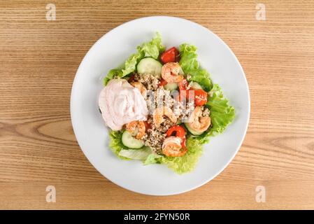 Salat mit Garnelen, Quinoa, Tomaten, Paprika, Gurken, Salat, Mayonnaise auf weißem runden Teller auf Holztisch. Stockfoto