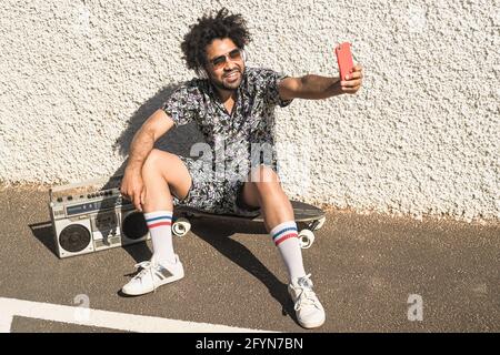 Junger afro-lateinischer Mann, der Spaß beim Selfie mit dem Handy hat Smartphone, während Sie Musik mit Kopfhörern und Vintage-Boombox hören Sommerferien Stockfoto