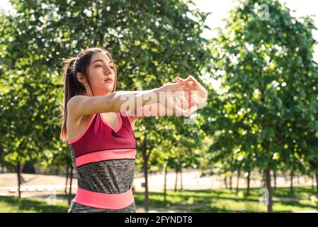 Horizontale Take einer schönen Frau, die ihre Arme streckt. Sie befindet sich in einem grünen Park und trägt rosa und graue Kleidung Stockfoto