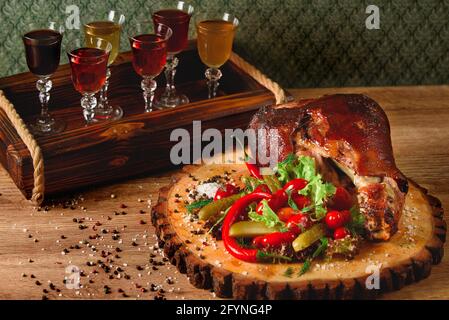 Schweinshaxe mit Gurken, Salat, Gemüse, Chili auf einem Holzbrett vor dem Hintergrund von Gläsern mit Alkohol. Stockfoto