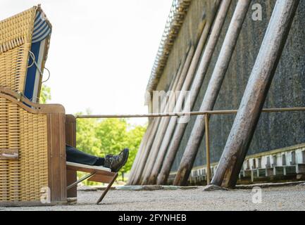 29. Mai 2021, Hessen, Bad Nauheim: Ein Mann hat es sich in einem Strandkorb vor dem Graduiertenhaus im Kurort gemütlich gemacht. Das Graduierungshaus besteht aus einem Holzrahmen, der mit Reisigbündeln gefüllt ist. Sole wird durch das Reisig geführt, was den Salzgehalt im Wasser erhöht und Wasser auf natürliche Weise verdampft. Gradierwerke, eigentlich Anlagen zur Salzgewinnung, werden heute in Deutschland oft zu Heilzwecken betrieben und sind daher besonders häufig in Kurorten anzutreffen. Die fallende Sole reichert die Luft in der Nähe der Graduierung an Stockfoto