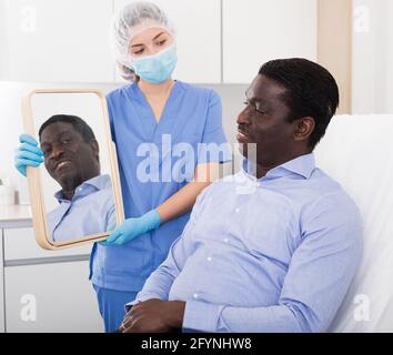 Glücklicher afroamerikanischer Mann, der den Spiegel ansah, während ihm eine professionelle Kosmetikerin Ergebnisse eines Hardware-Gesichtsverfahrens in einem medizinischen ästhetischen Arzt zeigte Stockfoto