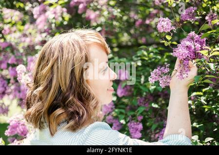 Ehrliches, authentisches Porträt einer kaukasischen blonden Frau aus den 30er Jahren mit Fliederblumen. 30 40-jährige Frau genießen das Leben in lila Blumen Natur Hintergrund Stockfoto