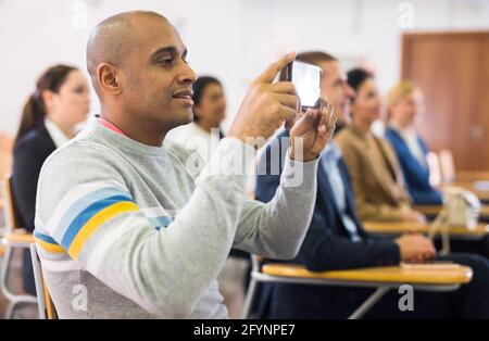 Porträt eines interessierten lateinamerikanischen Mannes, der an einer Geschäftsveranstaltung im Konferenzraum teilnimmt und mit dem Smartphone den Vortrag aufzeichnet Stockfoto