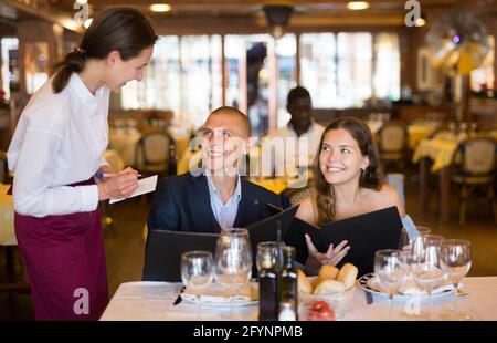 Freundliche, lächelnde Kellnerin, die ein paar Gäste bei der Auswahl von Getränken und Mahlzeiten im Restaurant beraten hat Stockfoto