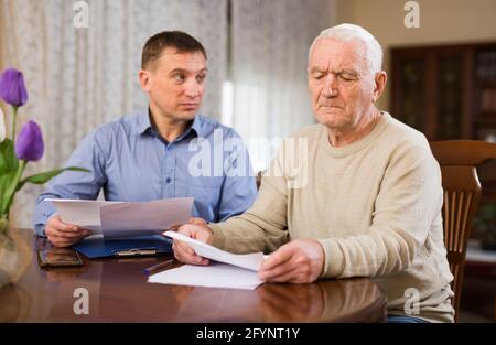 Beunruhigte ältere Mann diskutiert Dokumente mit seinem erwachsenen Sohn, während sie am Tisch zu Hause sitzen Stockfoto