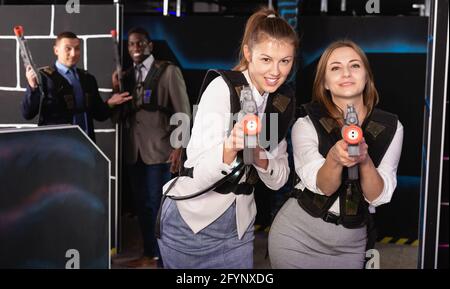 Emotionales Porträt von zwei fröhlichen, fröhlichen Frauen, die Laserpistolen halten und mit Kollegen Laser-Tag spielen Stockfoto