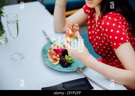 Mädchen in einem Café. Beschnittene Ansicht einer jungen Frau, die im Café im Freien Lachssandwich hält. Modische Mädchen tragen in roten Kleid. Stockfoto