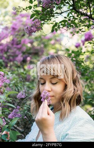 Ehrliches, authentisches Porträt einer kaukasischen blonden Frau aus den 30er Jahren mit Fliederblumen. 30 40-jährige Frau genießen das Leben in lila Blumen Natur Hintergrund Stockfoto